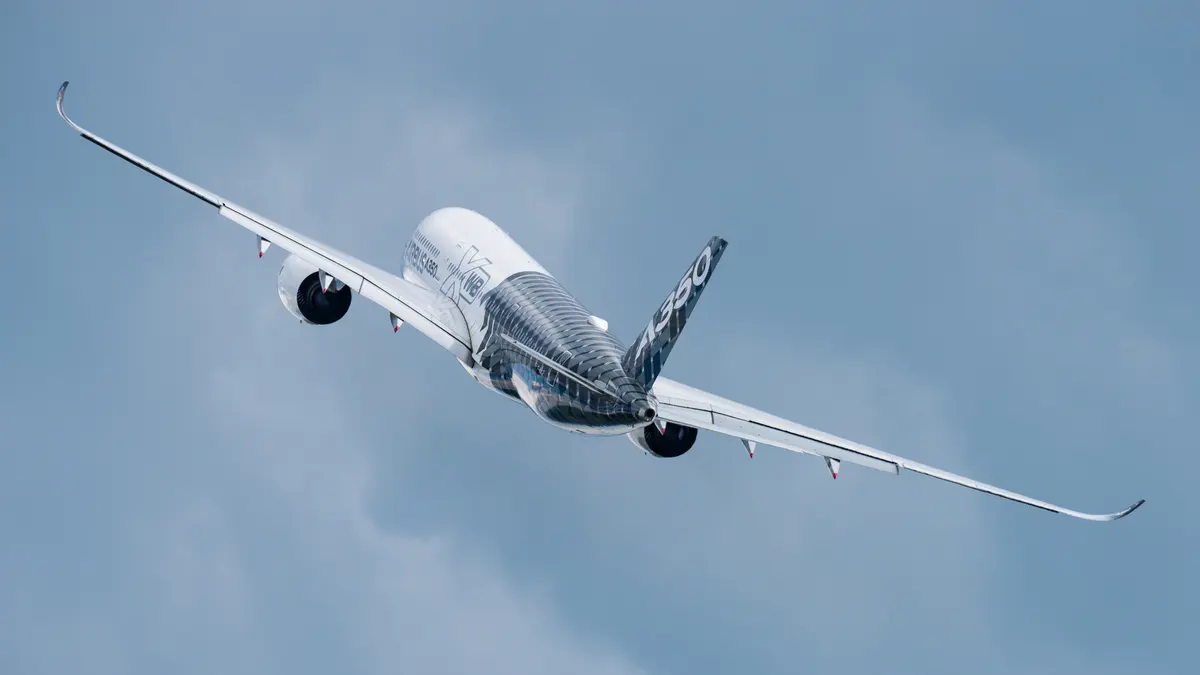An Airbus A350 banking in flight against a pale, hazy sky, seen from behind and to the side.