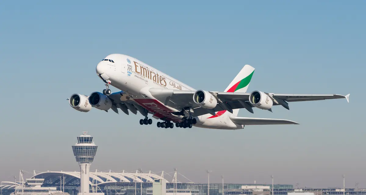 An Emirates Airbus A380 just airborne, with an airport control tower and terminal behind it.