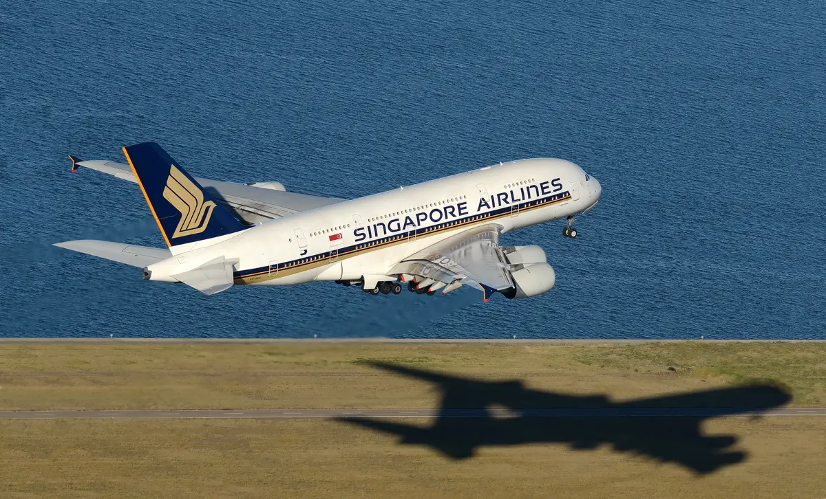 A Singapore Airlines Airbus A380 lifting off over water, its shadow stretching across the grass below.