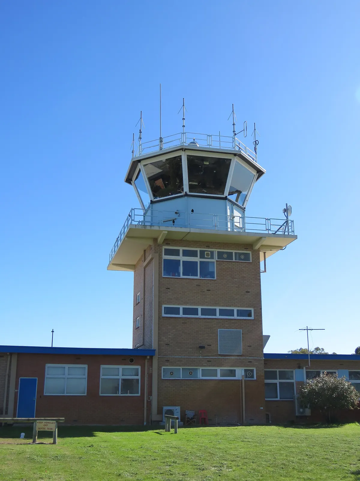 A small airport control tower with a glass cab and antennas, seen from the grass in front of it against a clear blue sky.