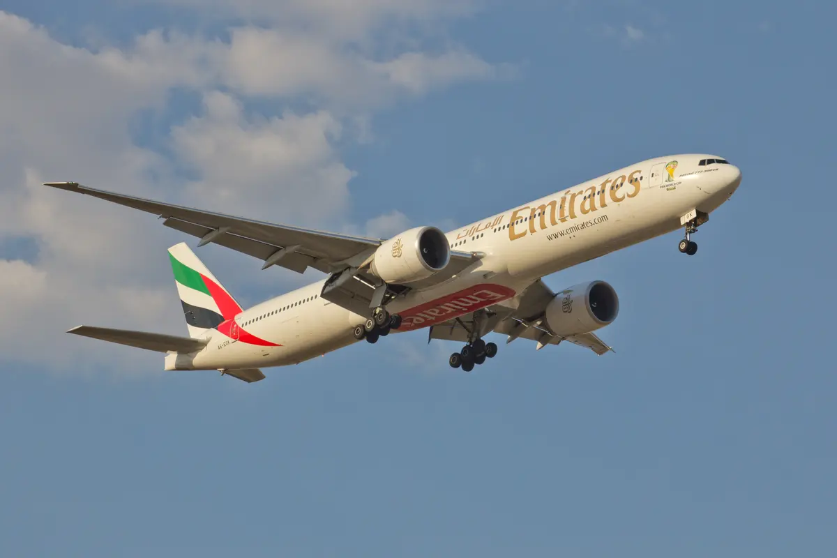 An Emirates Boeing 777-300ER in flight against a blue sky with a few clouds, gear retracting.
