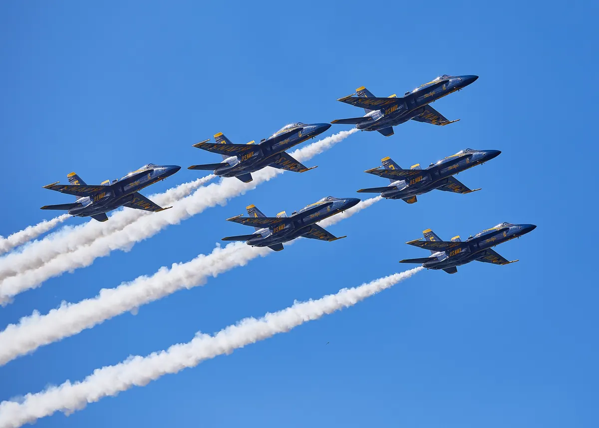 The Blue Angels flying their delta formation with white smoke trails across a clear blue sky.