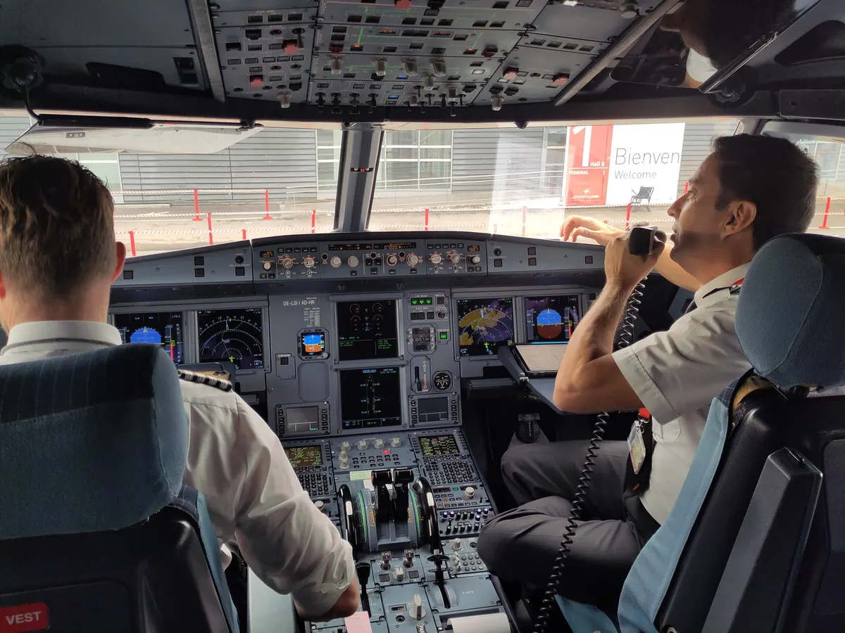 Two pilots in an Airbus A320 cockpit on the ground, one of them speaking on the radio, with the instrument panel and overhead panel around them.