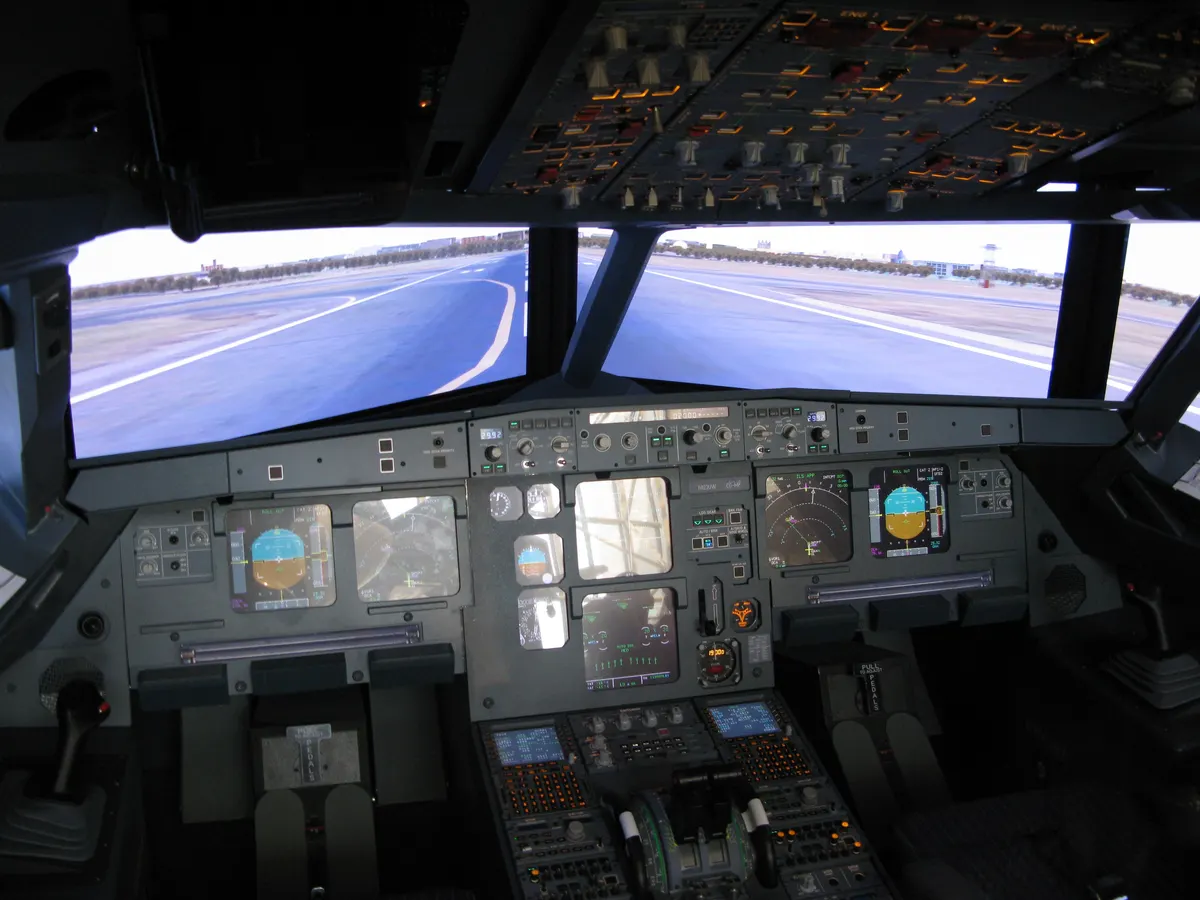The glass cockpit of an Airbus A320 seen from behind the seats, with the primary flight displays, the central pedestal and the overhead panel.