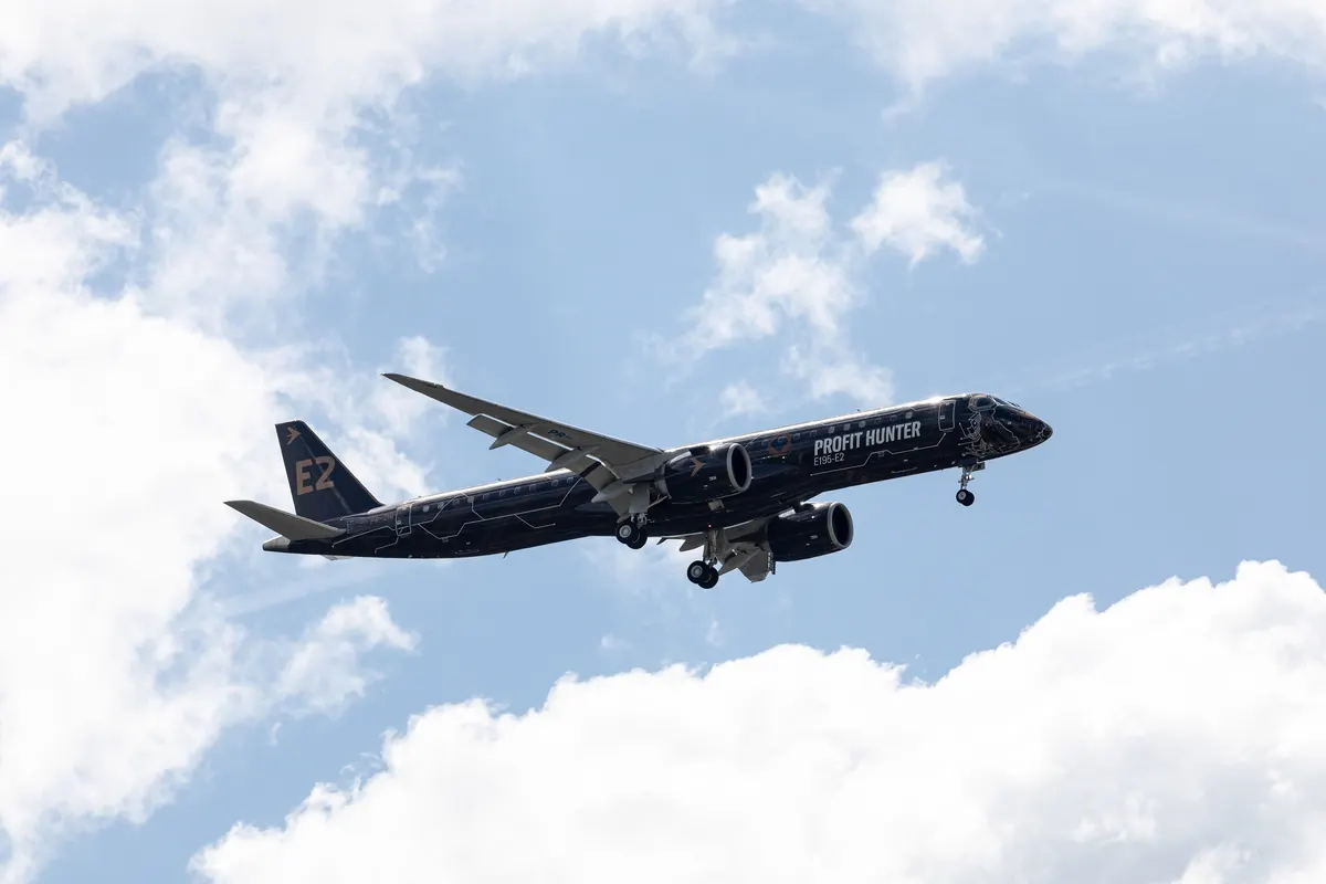 An Embraer E195-E2 in a dark livery banking in flight above white clouds at an air show.