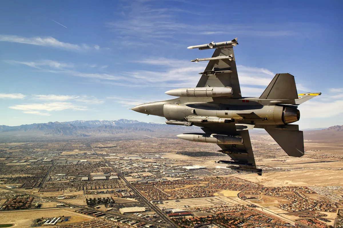 A USAF F-16 in a steep turn over the Las Vegas valley with mountains in the distance, seen from above with its wing pylons visible.