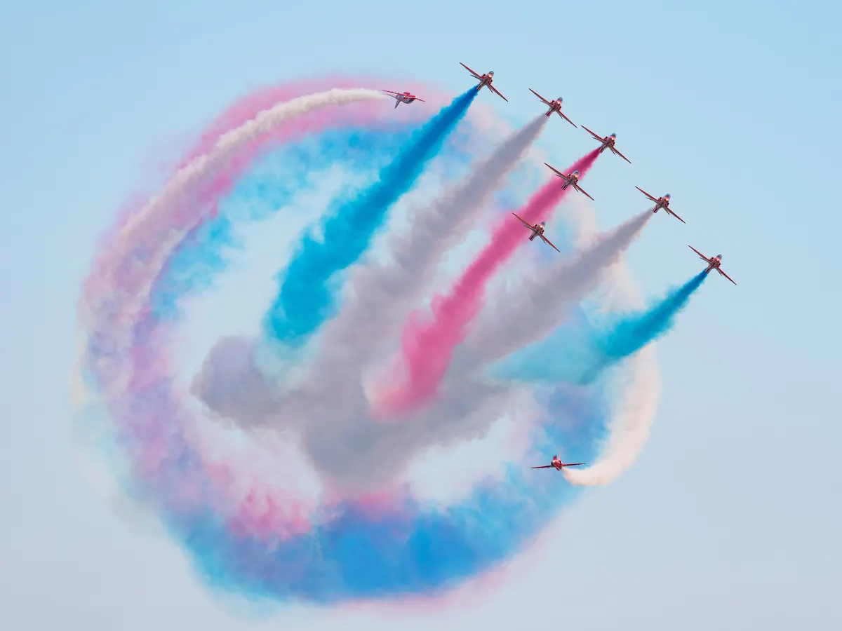 Seven Red Arrows jets looping through a circle of red, white and blue smoke against a pale sky.